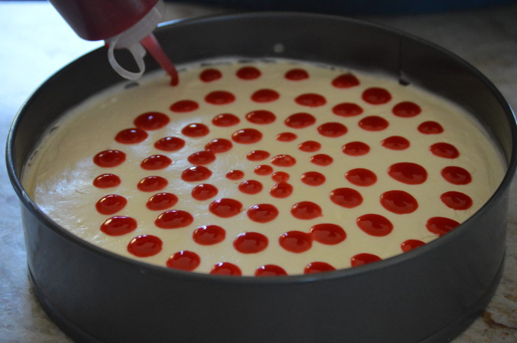 pouring out dots of raspberry sauce on top of the cheesecake